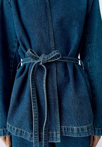 Veste en denim avec une taille cintrée et un nœud. Couleur bleu foncé, tissu texturé, couture visible et manches longues.