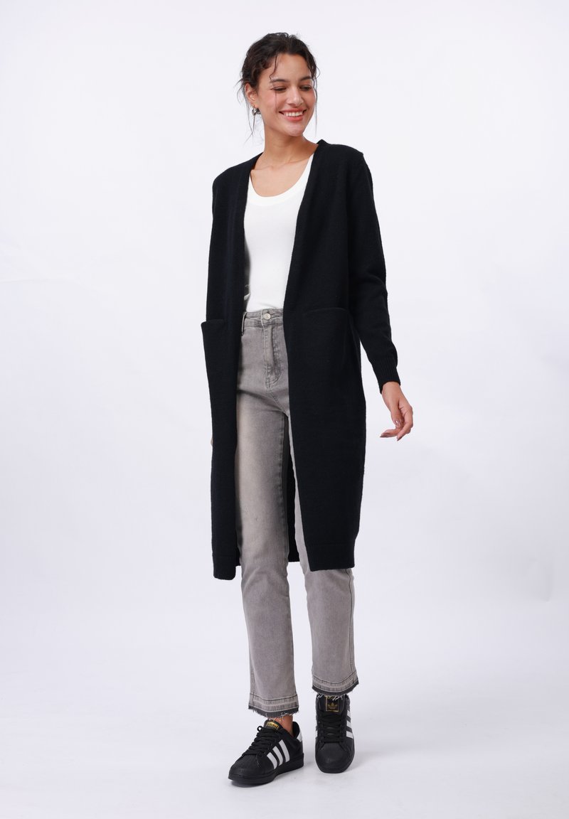 Black long cardigan with open front, two side pockets, and ribbed cuffs, paired with grey jeans and black sneakers. Smooth texture.