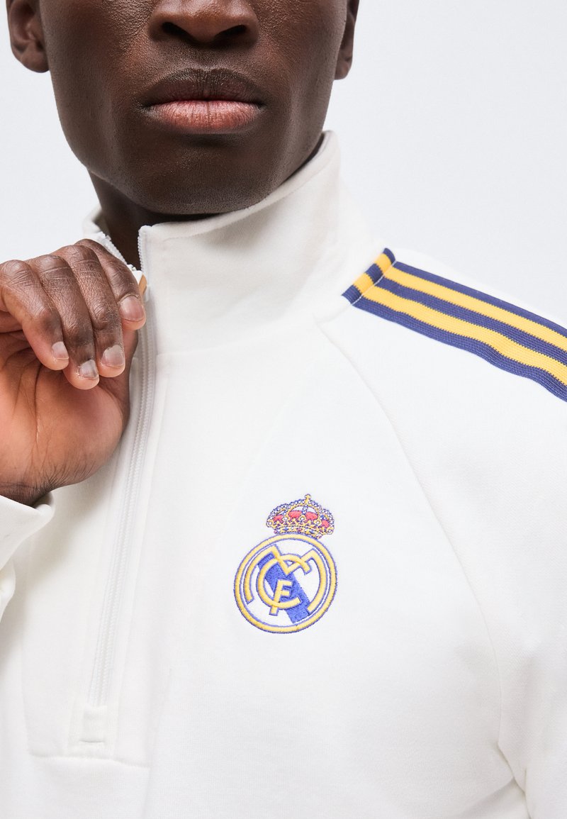 Gros plan d'une personne fermant une veste de sport blanche avec des rayures bleues et jaunes et un logo du Real Madrid sur la poitrine.