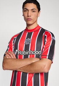 Mustade ja punaste triipudega lühikeste varrukatega jalgpallimärk, millel on kõrge kaelus, valge logo ja sponsoritekst. Sile tekstuur.