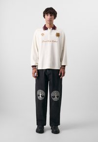 Witte lange mouwen polo met maroon kraag, met geborduurde tekst. Zwarte broek met ingewikkelde boomontwerpen op de knieën. Zwarte schoenen.