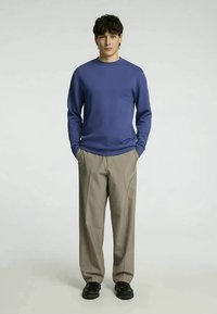 Σκούρο μπλε μακρυμάνικο πουκάμισο από βαμβάκι, συνδυασμένο με ανοιχτό καφέ παντελόνι και μαύρη υπόδηση, τοποθετημένο μπροστά από έναν μονόχρωμο φόντο.