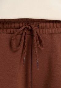 Bruine joggerpants met een geribbelde tailleband, voorzien van een trekkoordsluiting en zilverkleurige metalen tips aan de trekkoorden. Gladde textuur.