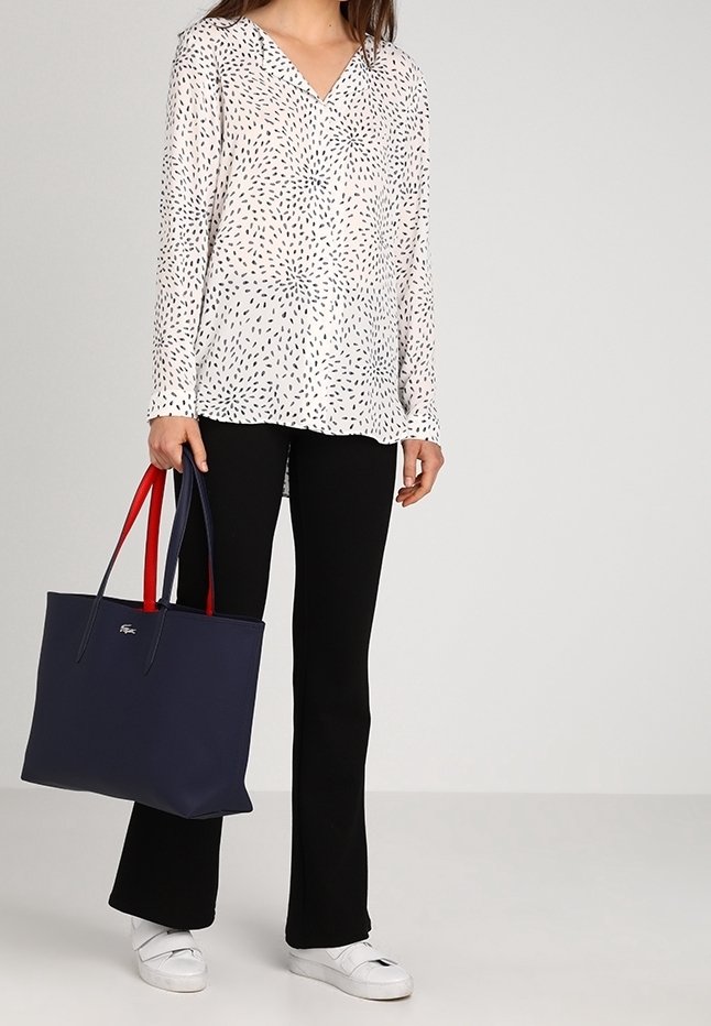 Femme portant un chemisier blanc à motifs, un pantalon noir et des baskets blanches, tenant un grand sac cabas bleu marine avec des anses rouges et bleues.