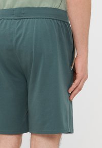 Calções masculinos em azul-petróleo escuro com cintura elástica; tecido macio, bolsos laterais e costuras limpas ao longo das bainhas. Projetados para conforto e facilidade de movimento.