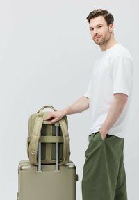 Olivgrön ryggsäck med vadderade remmar vilar på en silverfärgad resväska med hjul. Mannen står bredvid den, klädd i en vit t-shirt och gröna byxor.