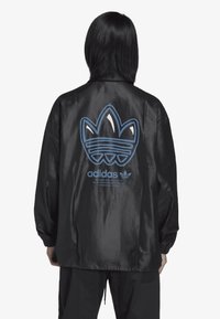 Veste noire à revêtement, arborant un grand logo trefoil Adidas bleu à l'arrière, avec des poignets élastiques et un design décontracté.