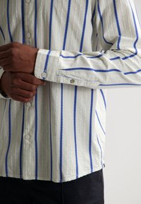 Weißes Hemd mit Knopfleiste, blauen vertikalen Streifen und strukturiertem Muster; mit Knöpfen und Manschetten-Details.