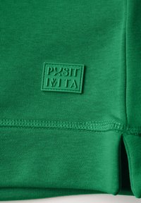 Grön textil med en mjuk, slät yta, med en sydd fåll och en upphöjd grön gummilapp som läser "POSITIVITA."