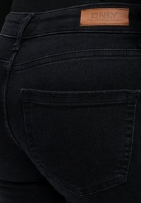 Gros plan de la poche arrière d'un jean en denim noir avec une étiquette en cuir marron portant la mention "ONLY Blue Denim anno 1995" sur la ceinture.