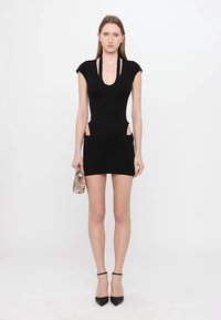 Rochie neagră, ajustată, cu mâneci scurte, tăieturi pe laterale și decolteu adânc. Modelul poartă o geantă mică cu un model și tocuri negre, ascuțite.