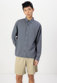 Camisa de botón gris, tejido texturizado, de mangas largas. Combinada con pantalones cortos de color beige claro, de cortes informales. Modelo lleva gafas de sol sobre la cabeza.