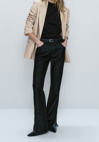 Pantalons en denim noir évasés à taille haute, associés à un haut noir ajusté et à un blazer beige. L'ensemble est complété par des talons aiguilles noirs.