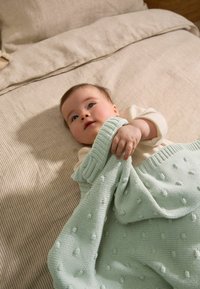 Couverture en tricot mint douce et texturée avec un motif en relief de pompons, tenue par un enfant sur un fond de lit beigné à rayures.