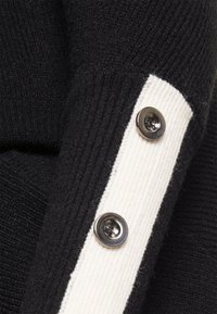 Dettaglio di maglione nero lavorato a maglia con una striscia verticale bianca e bottoni metallici come decorazione sulla manica. Tessuto strutturato con motivo a coste.