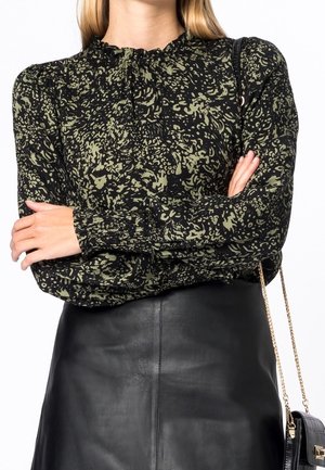 Mujer con los brazos cruzados que lleva un top de manga larga negro y verde con estampado y una falda de cuero negra, sosteniendo un bolso negro con una cadena dorada.