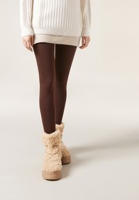 Brune leggings parret med en lang cremet ribbet sweater og beige fluffy ankelstøvler. Støvlerne har en tekstureret pelslignende overflade.