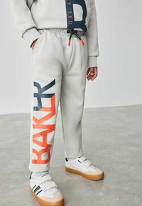 Hellgraue Sweatpants mit einem großen navyblauen und roten "BAK"-Grafik auf dem linken Bein, ausgestattet mit einem elastischen Bund und Kordelzug.
