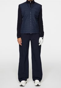 Marinblå pufferväst med vertikal sömnad, i kombination med löst sittande marinblå byxor. Har en zip-up-design och en golfhandske på höger hand.