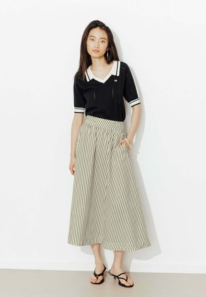 Femme portant un haut noir à manches courtes et col, ainsi qu'une jupe taille haute beige à fines rayures verticales noires, debout contre un mur blanc.