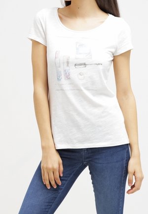 Kvinde iført hvid T-shirt med makeup-tema print og blå jeans, stående med den ene hånd hvilende på sit ben.