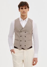 Beżowa kamizelka w kratę z podwójnym zapięciem, sześcioma ciemnymi guzikami i dwiema bocznymi kieszeniami, noszona na białej koszuli.
