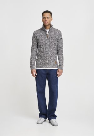 Suéter de punto gris y azul marino con cuello alto y cierre de media cremallera, combinado con pantalones azul oscuro y zapatillas gris claro.