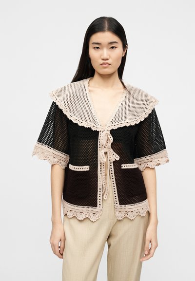 Νεαρή γυναίκα που φοράει μαύρο πλεκτό κοντομάνικο ζακέτα με μπεζ ανάγλυφη διακόσμηση και δέσιμο μπροστά, συνδυασμένο με μπεζ παντελόνι, στέκεται μπροστά σε λευκό φόντο.