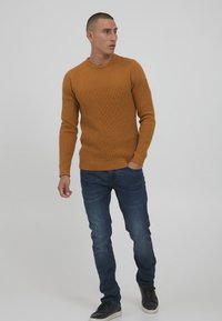 Brun strikkegenser med teksturert design, rund hals, lange ermer, brukt med mørkeblå jeans og svarte joggesko.