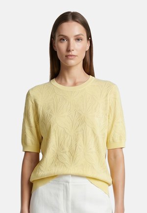 Femme aux cheveux bruns raides portant un top en maille texturée jaune clair à manches courtes et un pantalon blanc taille haute sur fond blanc.