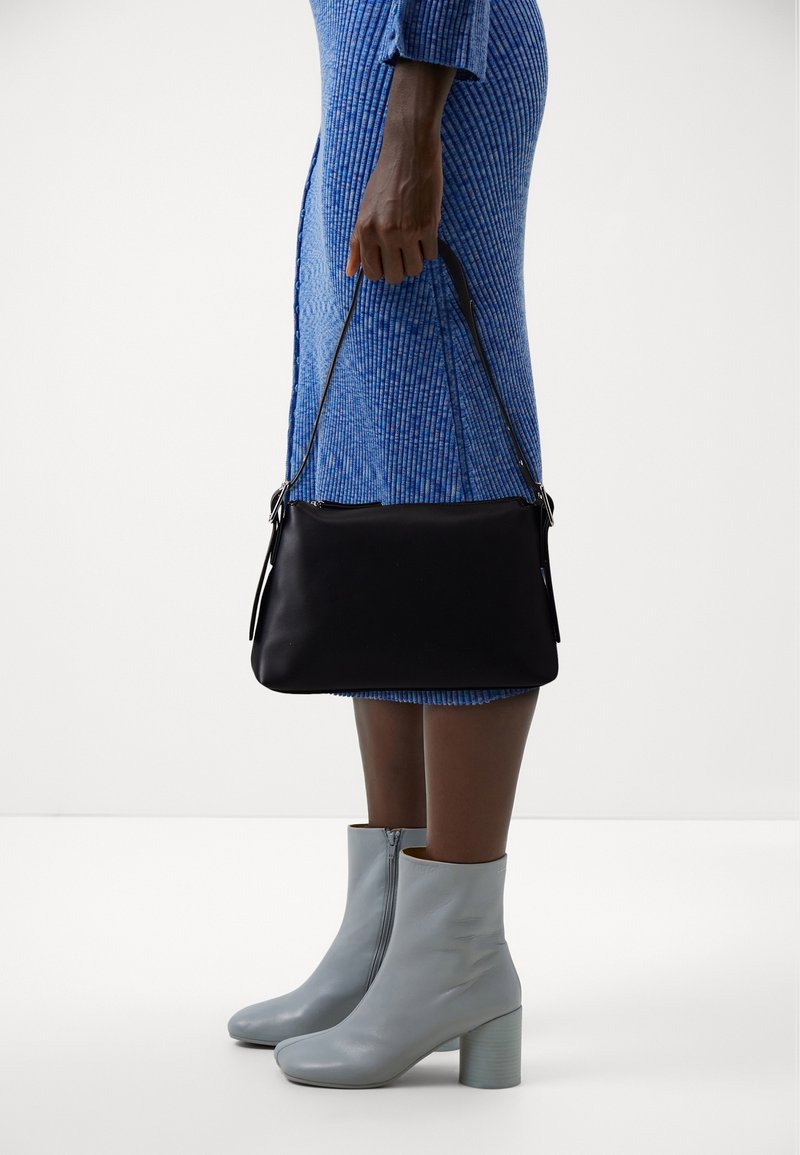 Personne en robe côtelée bleue tenant un sac à main noir, portant des bottines gris clair à talons blocs sur fond blanc.