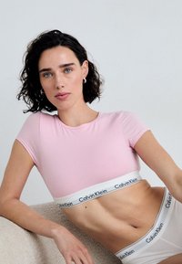 Světle růžový přiléhavý top s krátkými rukávy, spárovaný s bílým páskem Calvin Klein. Hladká textura, minimalistický design a čisté linie.