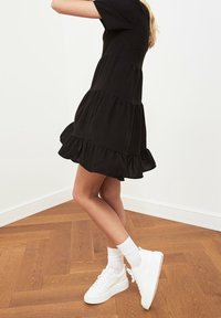 Robe noire à volants avec des manches courtes, en coton, ourlet à volants, associée à des baskets blanches à plateforme et des chaussettes mi-mollet. Fond en parquet.
