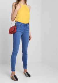 Top giallo senza maniche con dettagli in pizzo, jeans skinny blu con orli strappati e scarpe slip-on a fantasia bianco e nero, abbinate a una borsa rossa.