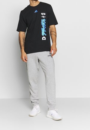 Camiseta de algodón negra con estampados gráficos en azul y blanco, combinada con pantalones deportivos grises que tienen franjas laterales negras y un logo de adidas.