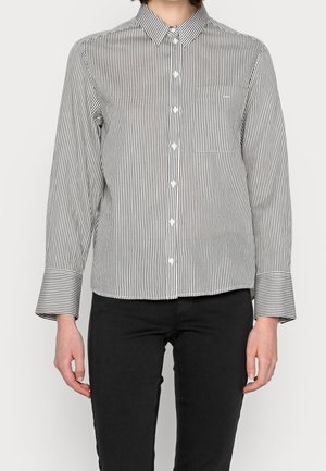 Donna che indossa una camicia button-up a righe verticali bianco e nero con maniche lunghe e pantaloni neri su uno sfondo semplice.