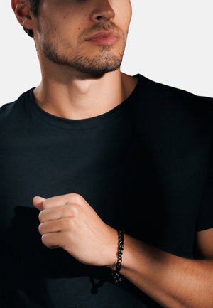 Svart kedjearmband på en handled, med sammanflätade länkar. Handen är delvis stängd, med en enkel svart t-shirt i bakgrunden.