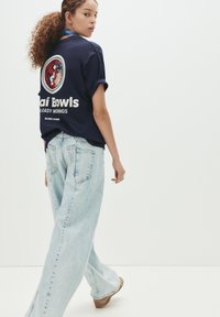 Marineblå bomulds T-shirt med "Açaí Bowls" grafik på ryggen, parret med lyseblå brede jeans, der har en falmet struktur og baglommer.