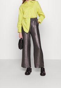 Limegrön skjorta med knappar, metallisk silverfärgade byxor med vida ben, svarta klumpiga klackstövlar och en liten svart handväska. Slät textur och avslappnad passform.