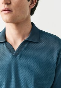 Polo de manga corta en verde azulado oscuro, con un patrón texturizado y un collar tipo V. La tela presenta un aspecto suave y ligeramente transpirable.