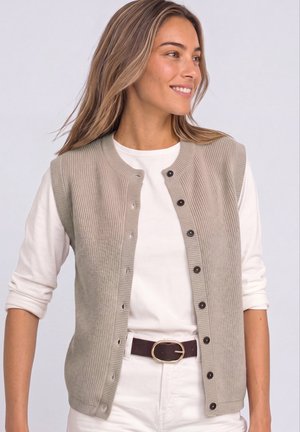 Femme aux longs cheveux souriante portant un gilet en maille beige boutonné par-dessus une chemise blanche à manches longues retroussées et un pantalon blanc avec une ceinture foncée.