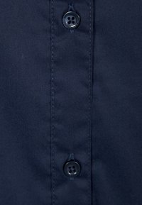 Mörkblått tyg med två sydda svarta knappar som är placerade vertikalt på en sydd ståndkrage.