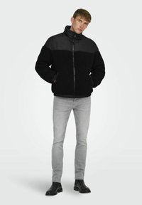 Chaqueta de forro polar negra con un panel negro liso en los hombros, cierre de cremallera y bolsillos frontales. Se lleva con jeans grises y botas negras.