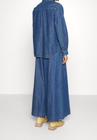 Henkilö, jolla on päällään löysä, sininen farkkupaita ja pitkä farkkuhame, yhdistettynä kiiltäviin vaaleankeltaisiin nauhallisiin kenkiin, seisoo harmaalla lattialla.