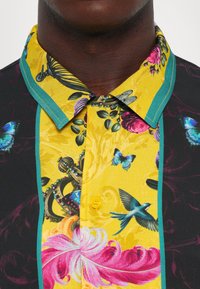 Cămășă neagră cu un panou galben, având un design cu păsări colorate, fluturi, flori și o coroană împodobită, purtată de o persoană cu pielea închisă la culoare.
