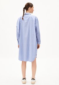 Robe chemise rayée en bleu clair et blanc, présentant une silhouette longue, un col boutonné et des fentes sur les côtés. Assortie avec des baskets décontractées.