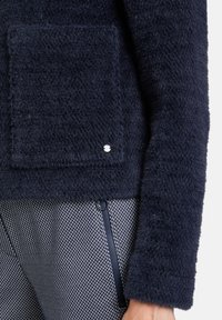 Granatowy, puszysty sweter z przednią kieszenią i srebrnymi akcentami. W połączeniu z fakturowanymi spodniami w czarno-białe wzory z bocznym zapięciem.