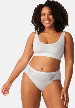 Lächelnde Frau mit lockigem Haar, die einen weißen nahtlosen BH und Spitzen-Slips trägt, steht vor einem schlichten hellen Hintergrund.