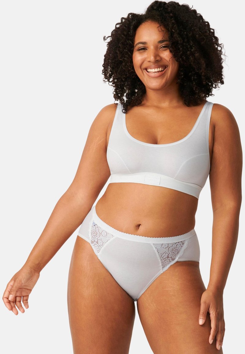 Femme souriante aux cheveux bouclés portant un soutien-gorge blanc sans couture et une culotte avec détails en dentelle, debout devant un fond clair uni.