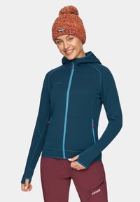 Turkusowa, zapinana na zamek kurtka z polaru z kapturem, o teksturowanej tkaninie i różowymi akcentami. Połączona z wielokolorową dzianinową czapką z pomponem.